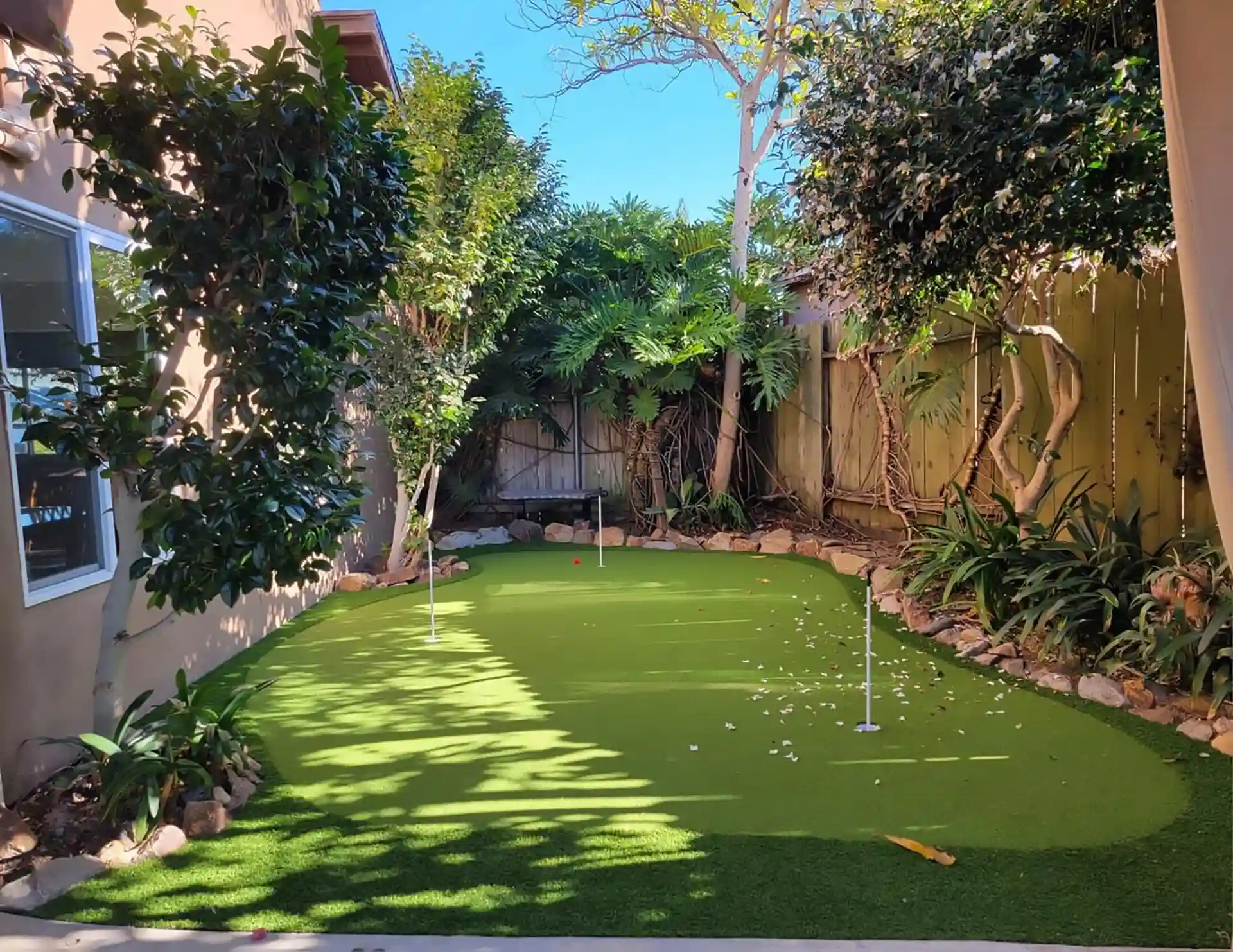 Photo of backyard putting green made of synthetic turf surrounded by lush plants and shaded by trees.

